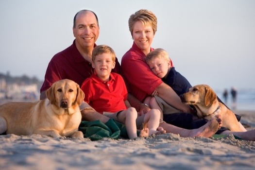 Ross, Mary, and their family enjoying the sunset at the beach, sitting together on the sand with the ocean in the background